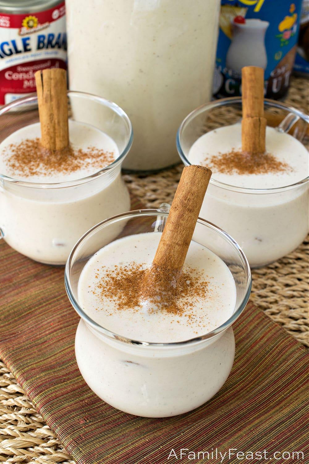 Top-down view of Coquito ingredients on a rustic wooden table: cans of coconut milk, evaporated milk, condensed milk, cream of coconut, bottle of white rum, cinnamon sticks, vanilla extract.