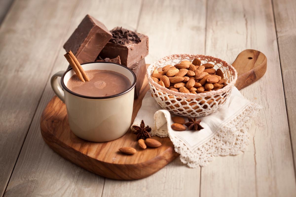 ingredients for almond milk hot chocolate laid out on a wooden table, focus on cocoa powder and almond milk carton