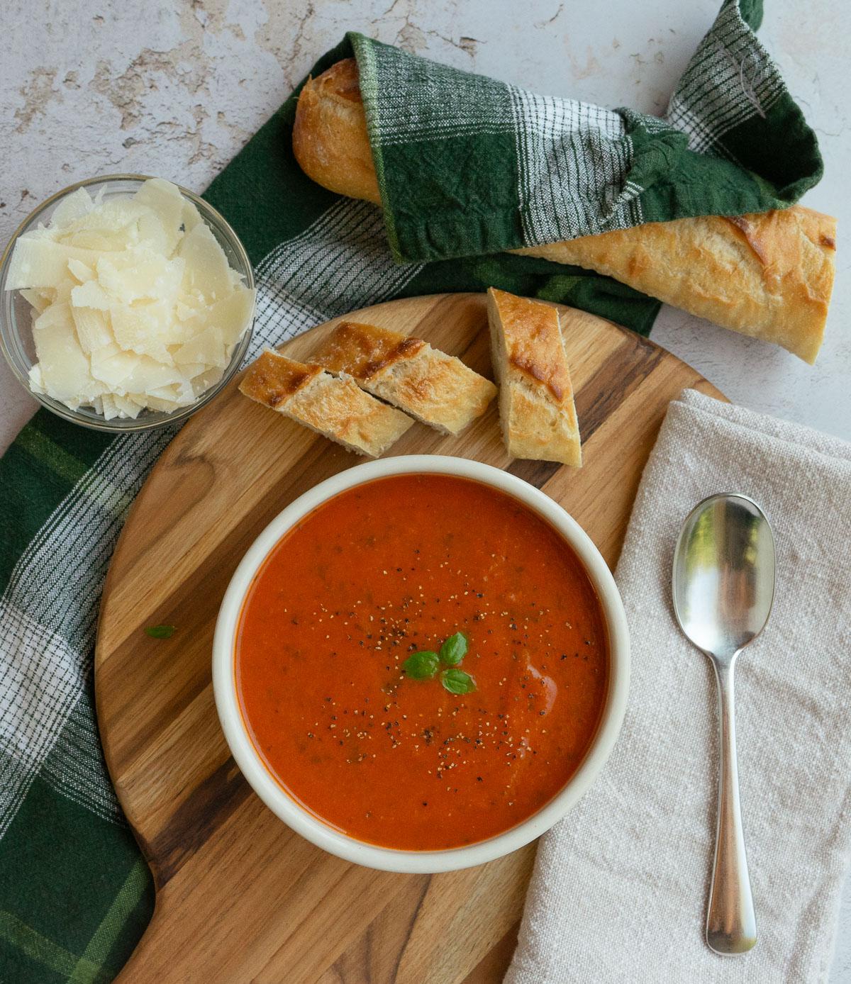 Overhead shot of a rustic table setting with bowls of tomato soup, a whole loaf of crusty bread, and fresh basil garnish