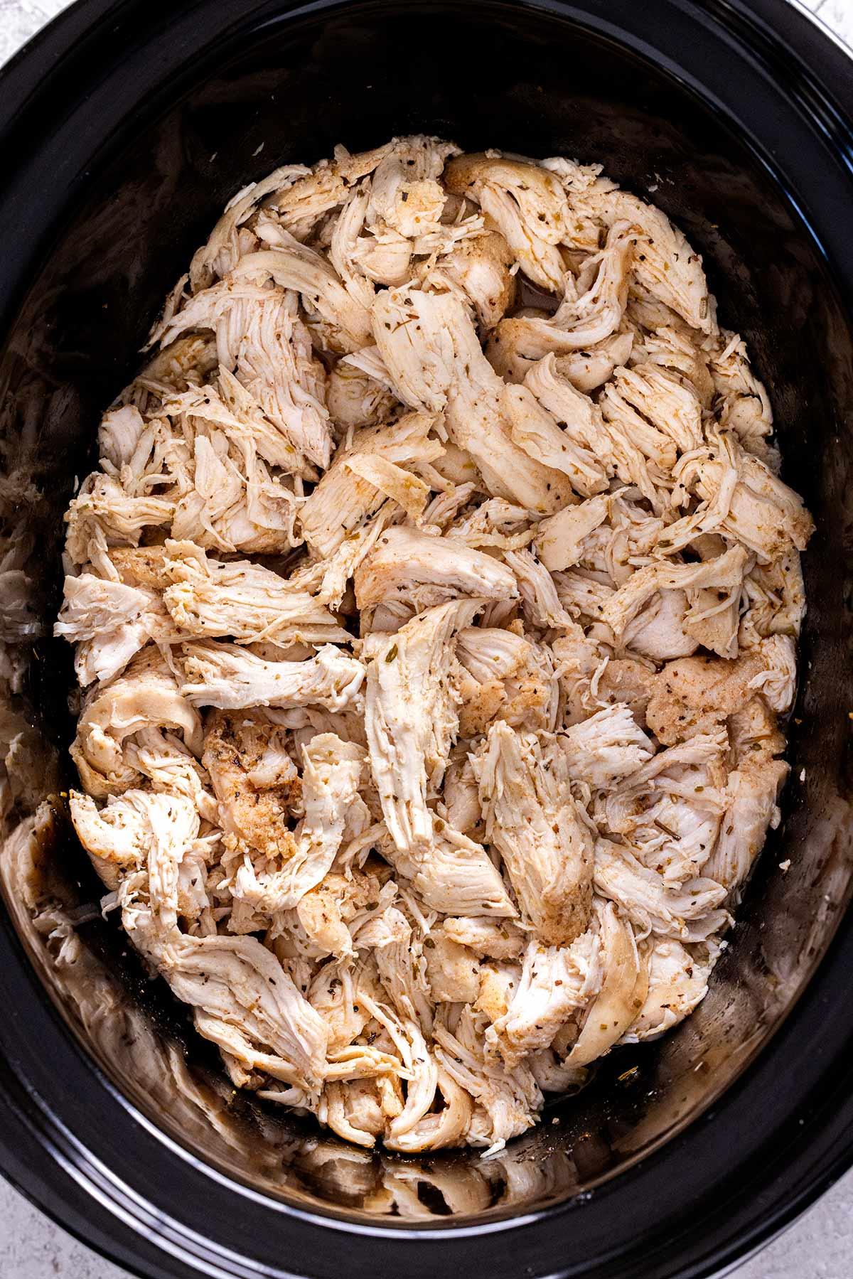 A close-up shot of shredded chicken in a crockpot, with herbs and spices visible