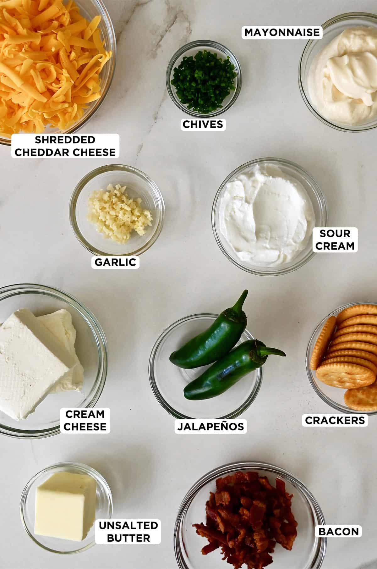 Ingredients for jalapeño popper dip laid out artfully on a light wooden cutting board: blocks of cream cheese, shredded cheddar, Monterey Jack cheese, fresh whole jalapeños, mayonnaise, and a bowl of sour cream.