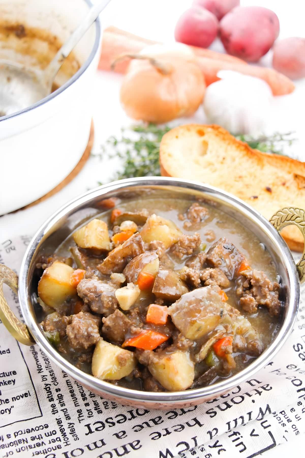 Ingredients for rustic beef ground stew laid out on a wooden cutting board: ground beef, carrots, potatoes, celery, onions, beef broth, herbs