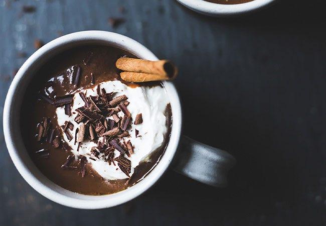 overhead shot of ingredients for spiced hot chocolate laid out on a dark surface: cocoa powder, sugar, cinnamon sticks, whole cloves, milk, vanilla extract