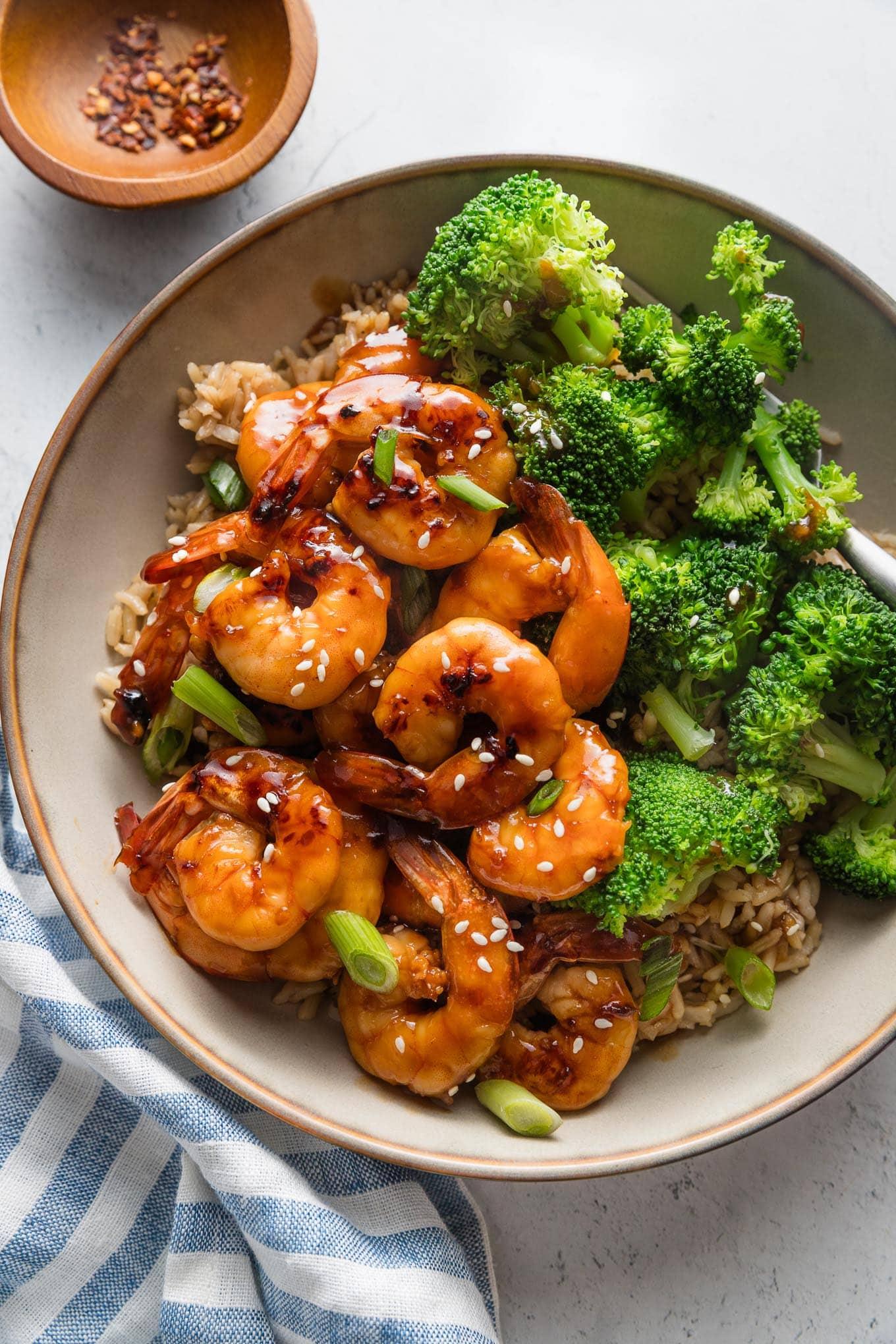 close-up shot of golden honey garlic shrimp in a hot pan, garnished with fresh chives, with rice and broccoli in the background