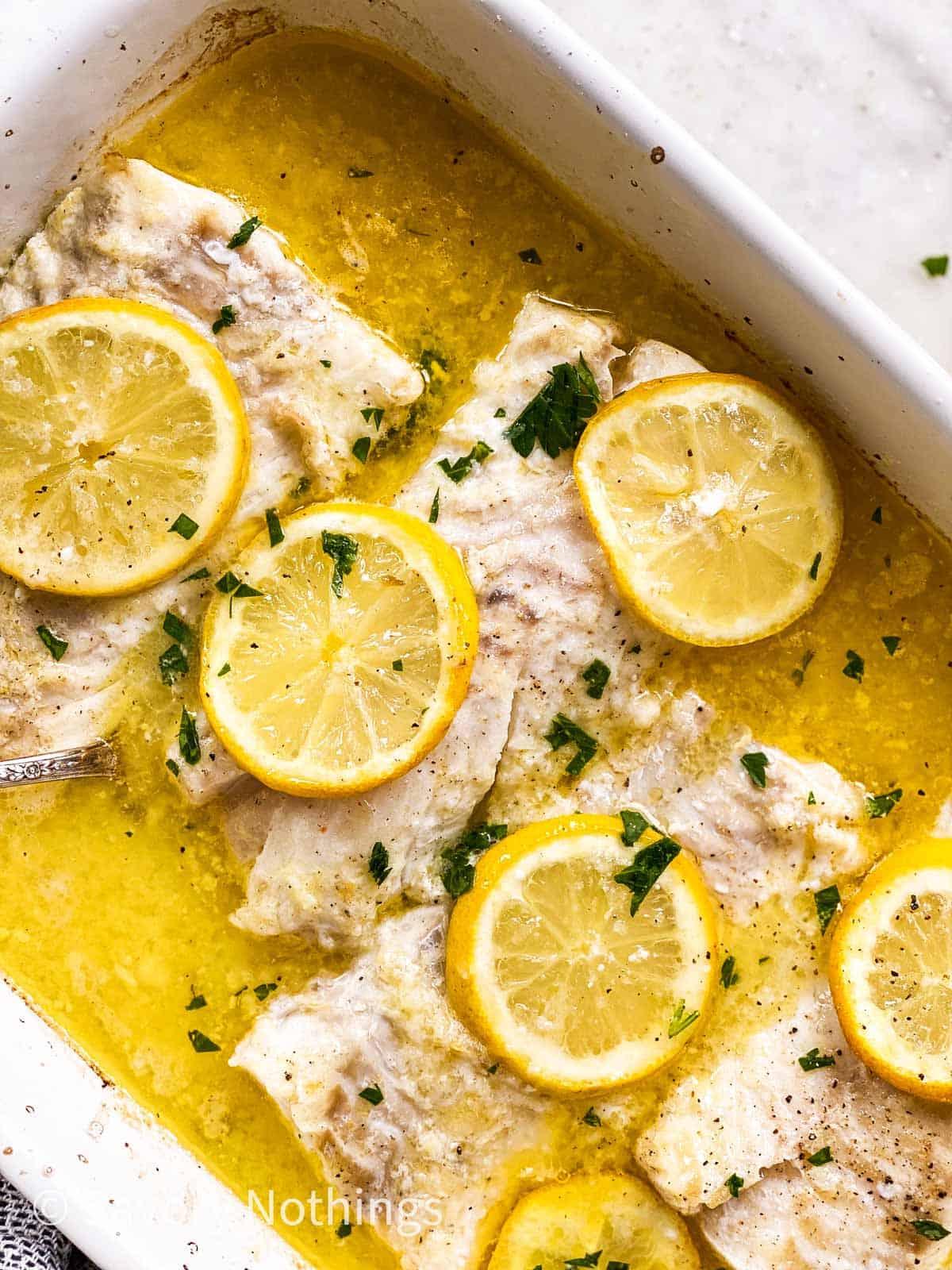 Hand drizzling lemon butter sauce over baked cod in a baking dish.