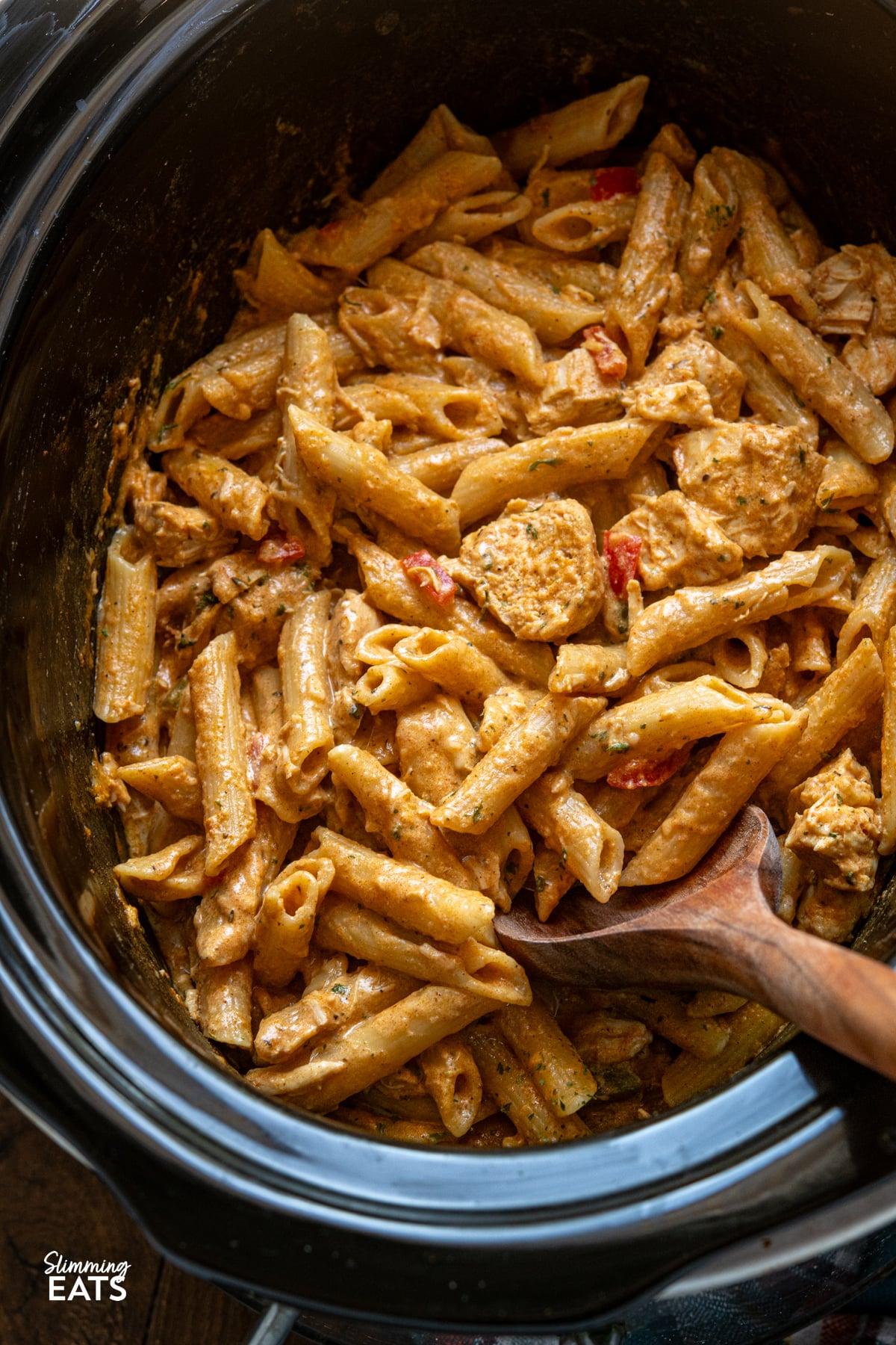 Close-up of cooked penne pasta being stirred into the creamy Cajun chicken mixture in a slow cooker