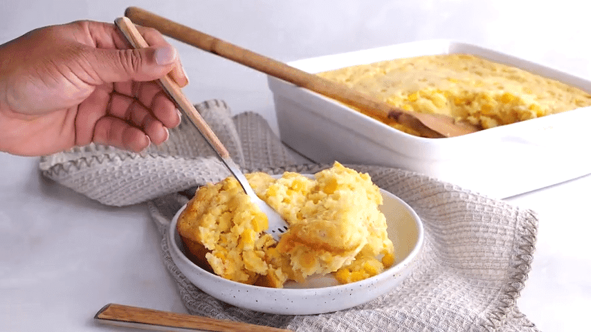 Close-up shot of a spoon scooping a fluffy, creamy portion of corn casserole from a baking dish, highlighting the texture.