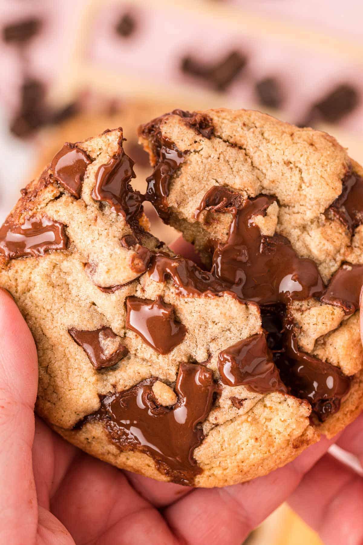 Hand breaking a chewy brown butter cookie in half, showing the soft interior and melted chocolate chips