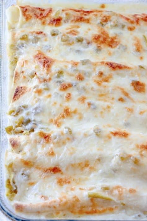 Close-up of creamy white sauce being poured over chicken-filled tortillas in a baking dish before baking.