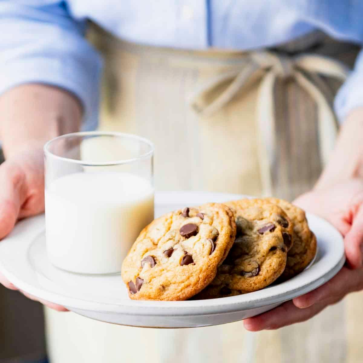 Hand reaching for a warm chocolate chip cookie on a cooling rack with a glass of milk