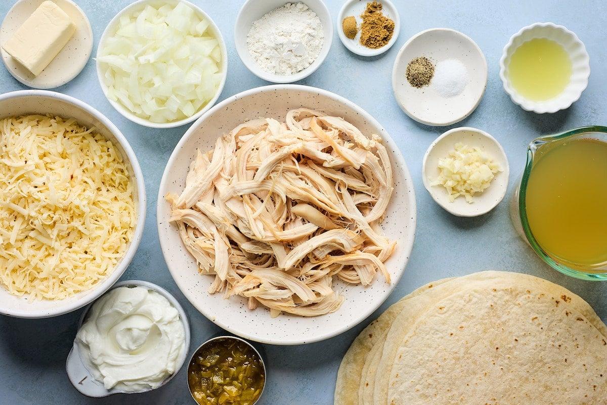 Overhead shot of all fresh ingredients for white chicken enchiladas laid out on a wooden table, including shredded chicken, tortillas, cheese, and cream.