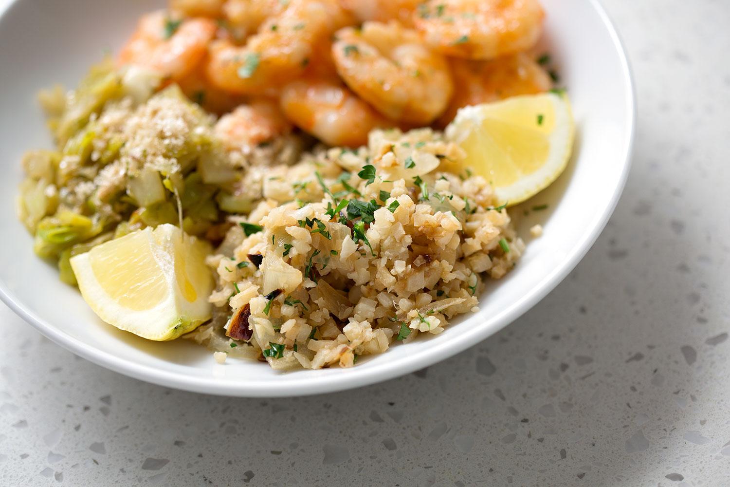 Overhead shot of fresh ingredients for cauliflower rice pilaf on a wooden cutting board: cauliflower, herbs, garlic, lemon