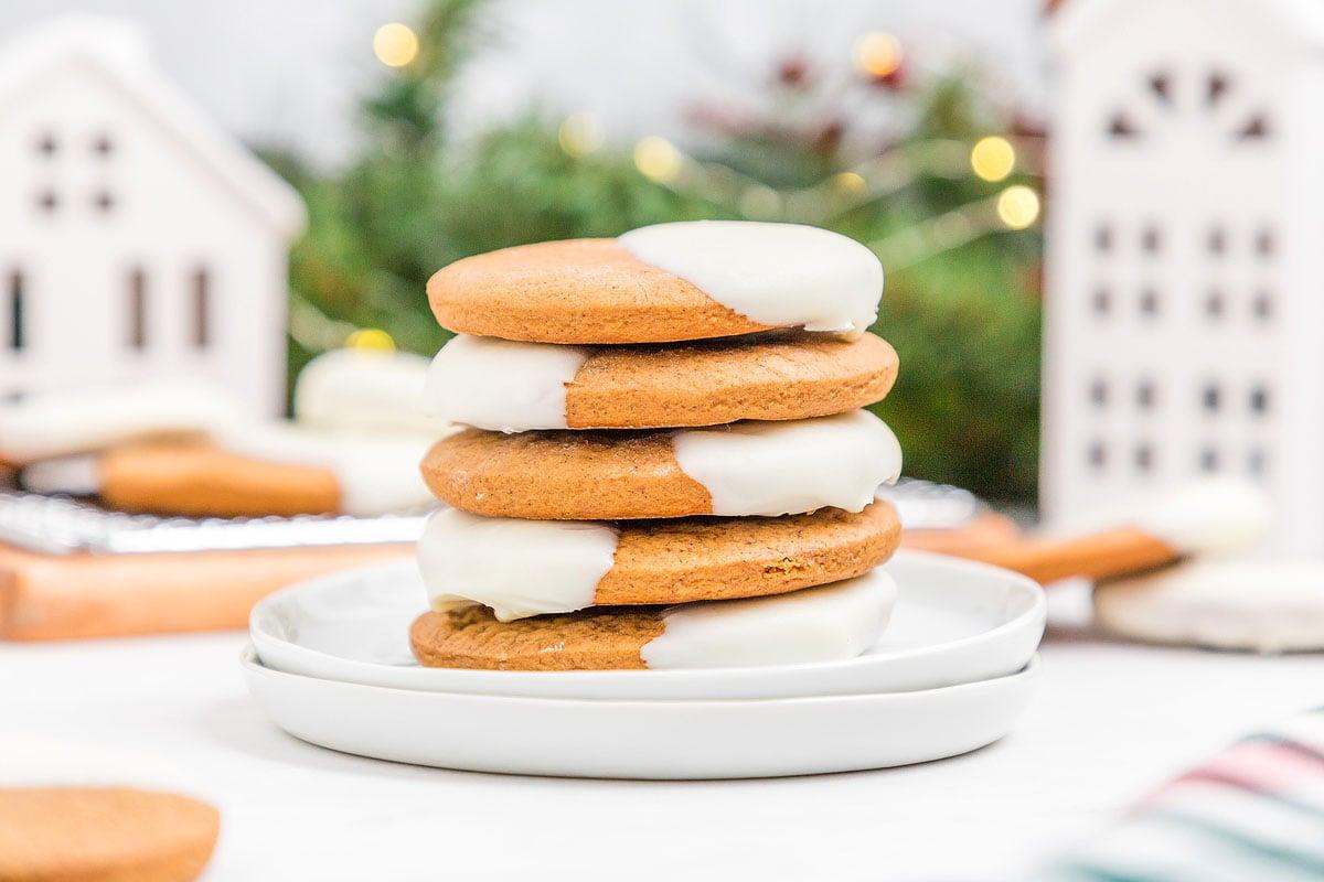 A stack of white chocolate dipped gingerbread cookies on a rustic wooden board with festive lights in the background.