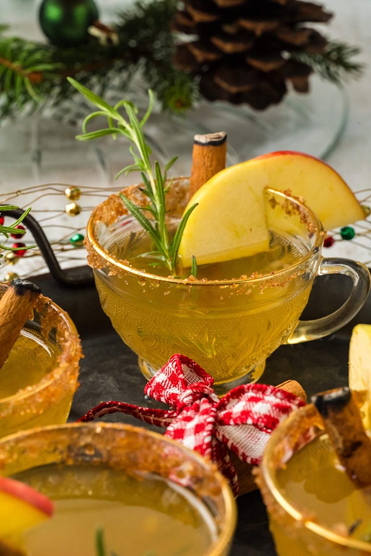 Pouring festive spiced apple drink into clear mugs, garnished with apple slices and cinnamon sticks