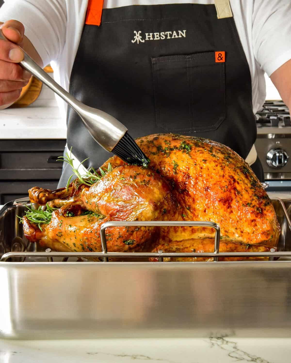 a person carefully basting a large roasted turkey in a deep roasting pan with a basting brush, showing golden drippings at the bottom of the pan