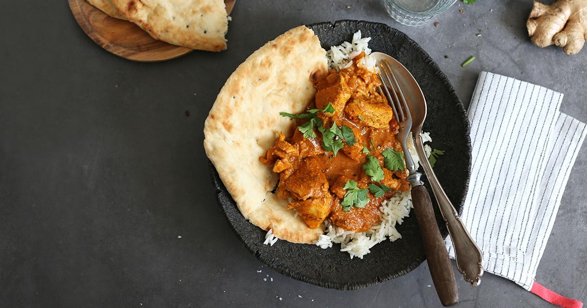 rustic overhead shot of a pot of rich, creamy butter chicken with naan bread and basmati rice on the side, steam rising