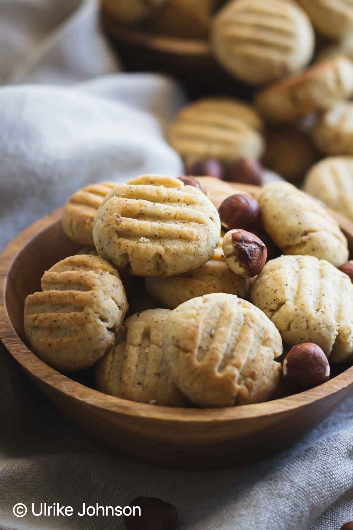 Close-up of golden brown hazelnut cookies stacked on a wooden board, with scattered whole hazelnuts in the background.
