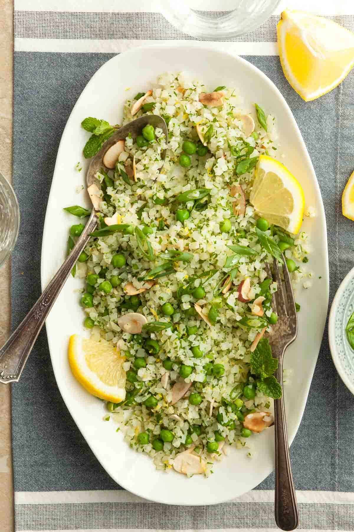 Close-up of a vibrant cauliflower rice pilaf with fresh herbs, garnished with lemon wedges, in a rustic bowl on a wooden table.