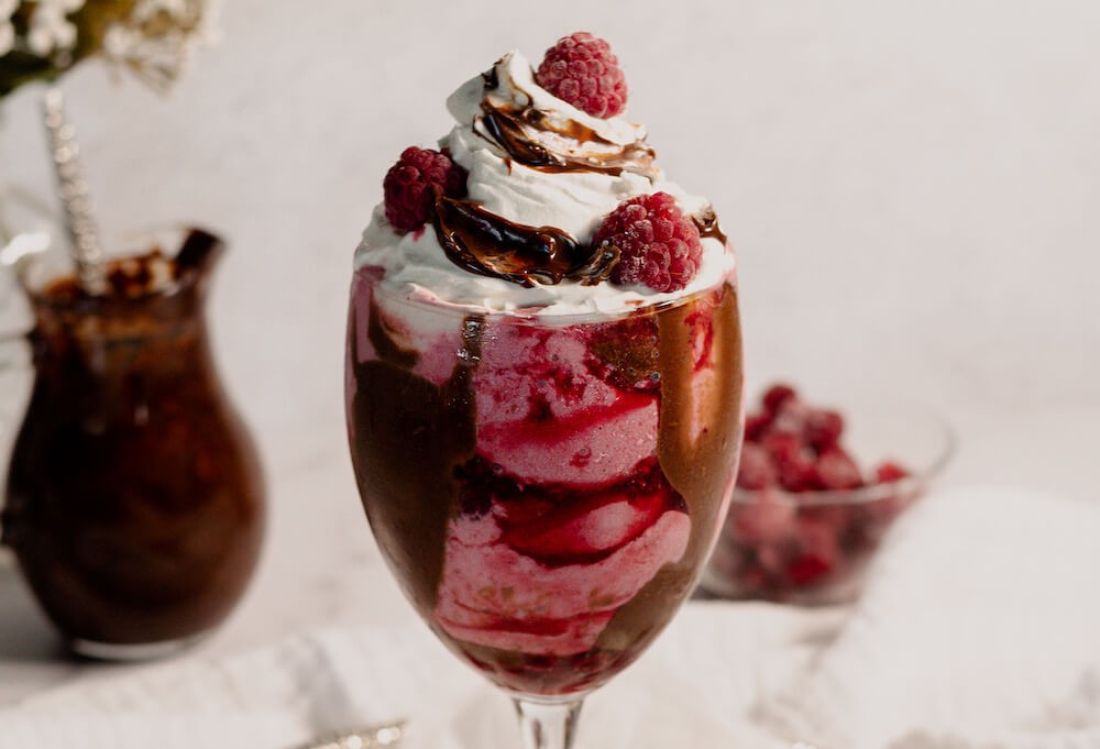 a vibrant, layered milkshake with swirls of milk, chocolate chunks, marshmallows, and raspberries, topped with whipped cream and a cherry