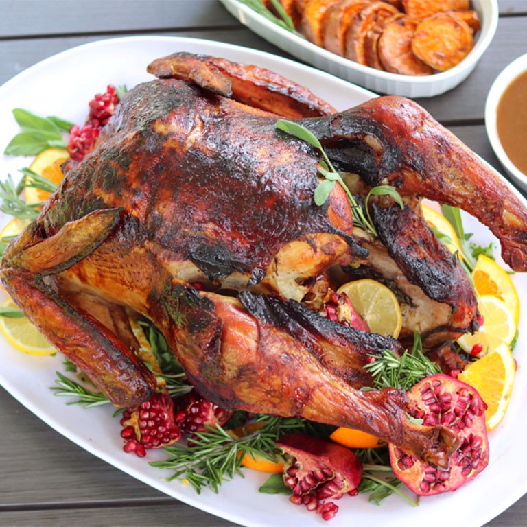 whole roasted brown butter sage turkey, golden crispy skin, surrounded by fresh herbs like sage and thyme, with a small ceramic pot of rich pan drippings next to it, set on a rustic wooden table with soft, festive lighting in the background