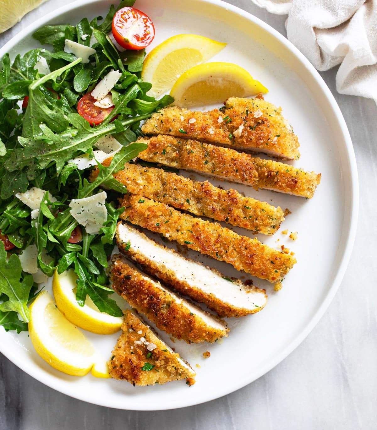 Crispy golden-brown parmesan chicken cutlets on a plate with fresh parsley, lemon wedges, and a light salad.