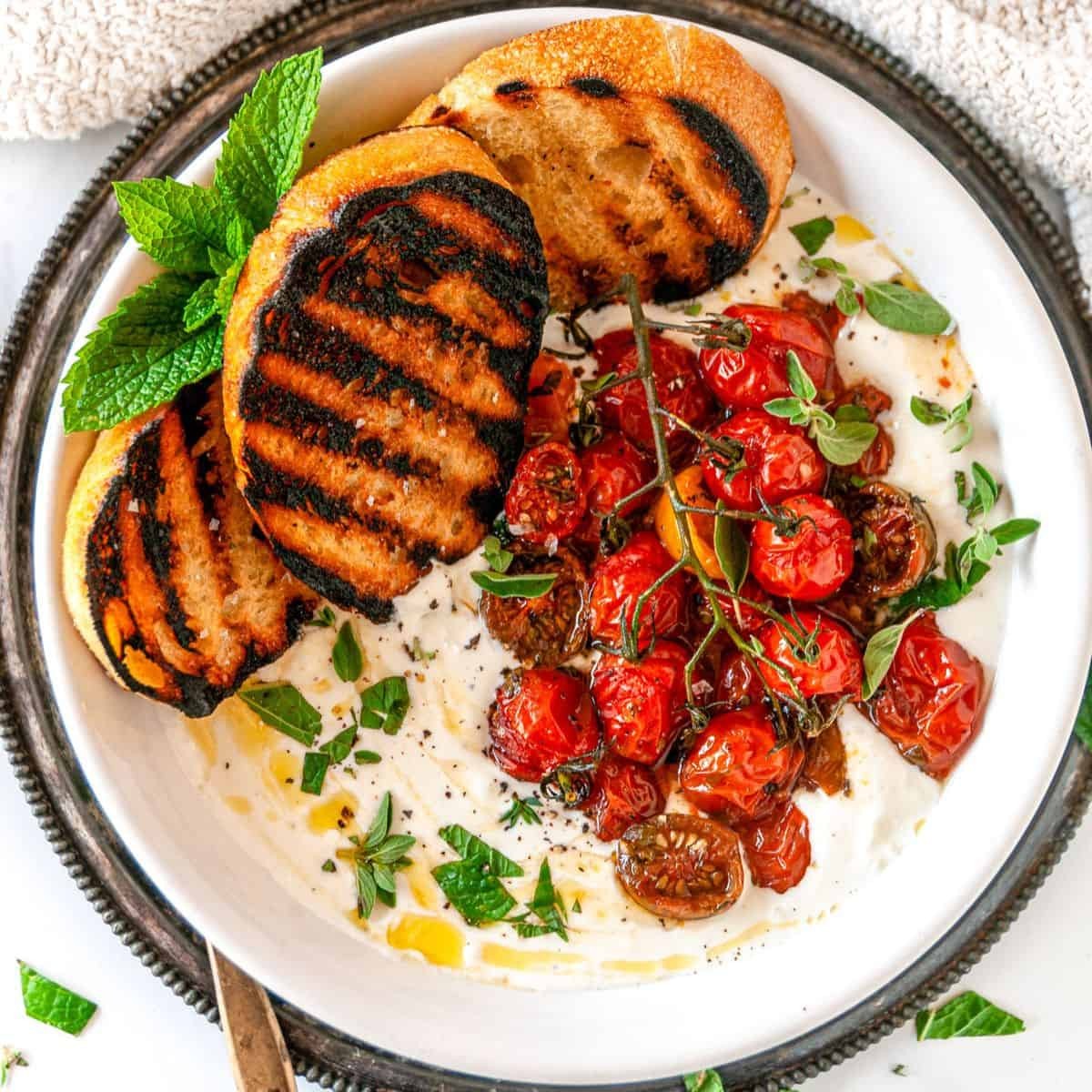 Vibrant whipped feta dip with roasted cherry tomatoes and fresh basil on a rustic wooden board, garnished with olive oil and cracked black pepper, next to crusty bread and pita chips, natural light