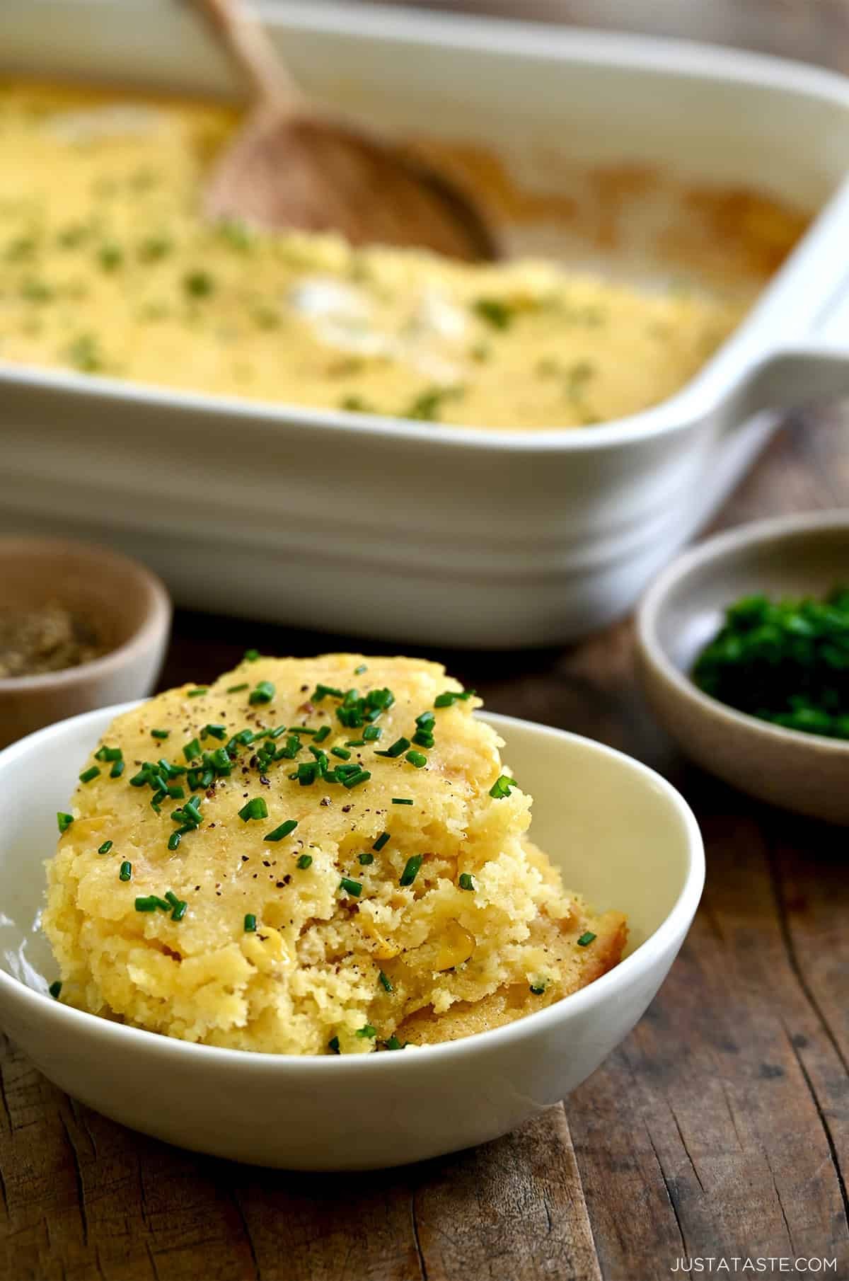 Close-up of a golden brown corn casserole with a visible melted butter center, garnished with fresh chives, rustic kitchen setting